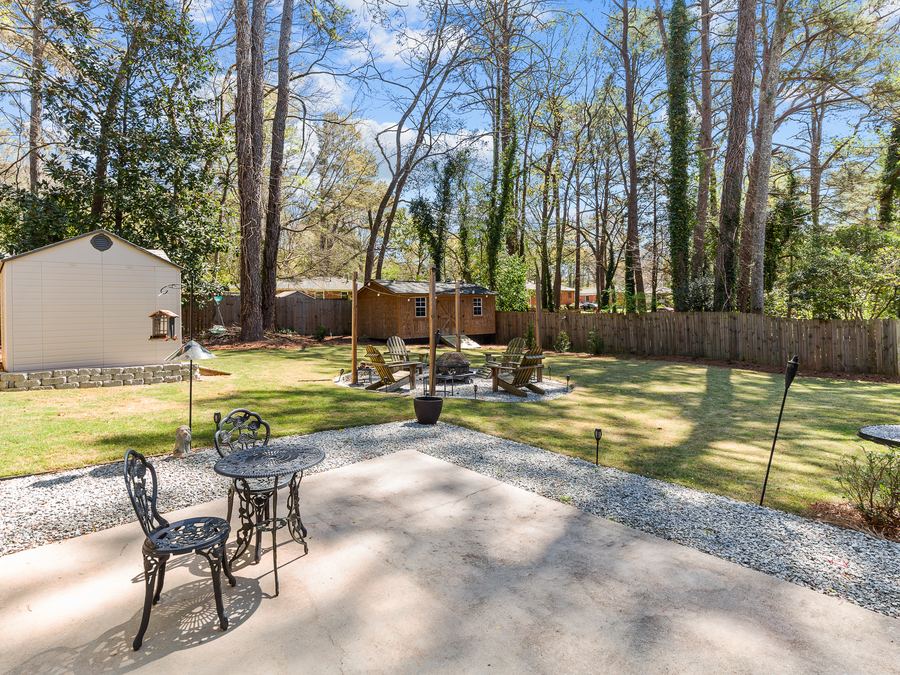 A shaded backyard patio with a bistro table, firepit seating and mature trees